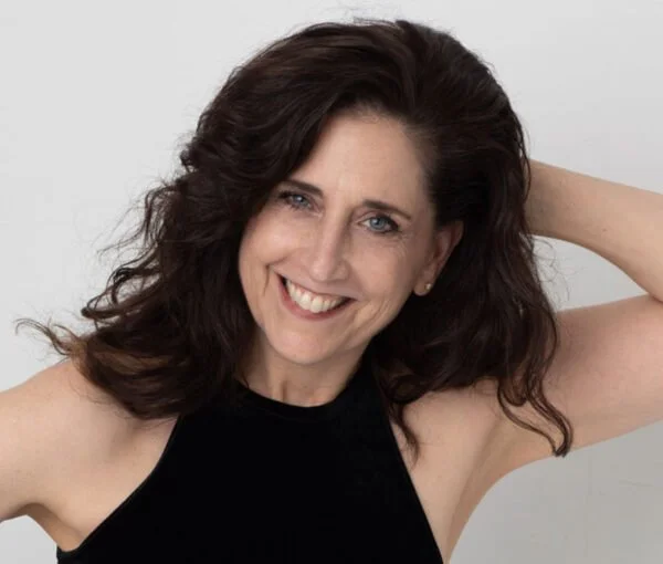 A woman with dark, wavy hair smiling and touching her hair, wearing a black sleeveless top, against a plain light background.