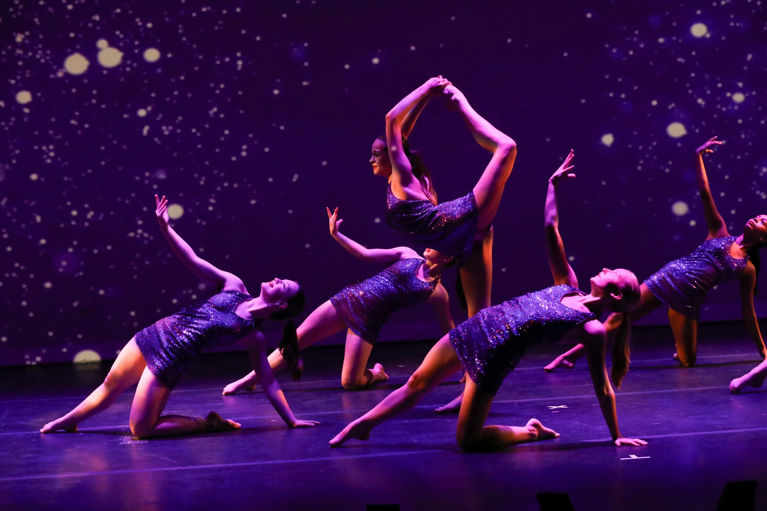 Group of dancers in glittery purple costumes performing a contemporary dance routine on stage with a starry background projection.