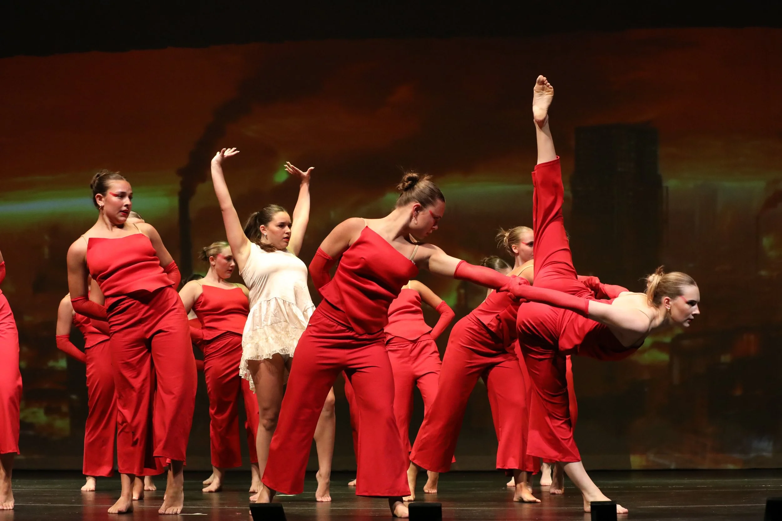 Group of women dancers in red costumes performing on stage with a cityscape and smoke in the background.