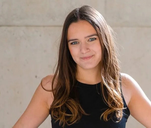 A young woman with long brown hair and blue eyes, wearing a sleeveless black top, smiling slightly in front of a plain light-colored wall.