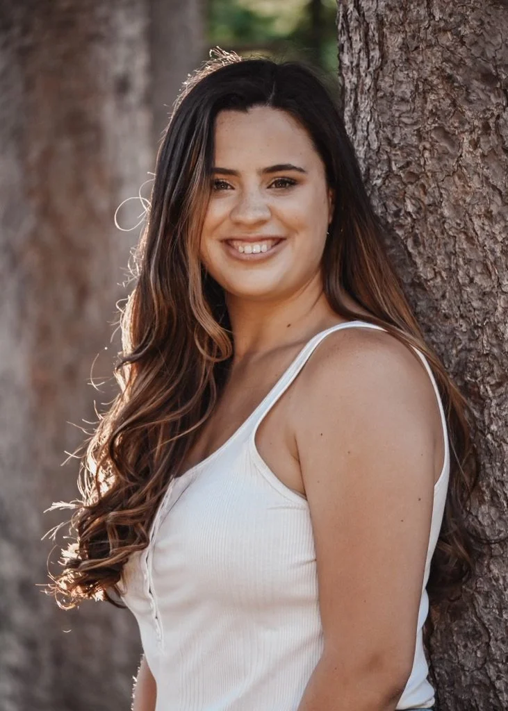 Young woman with long, wavy brown hair smiling outdoors, leaning against a tree, wearing a white sleeveless top.