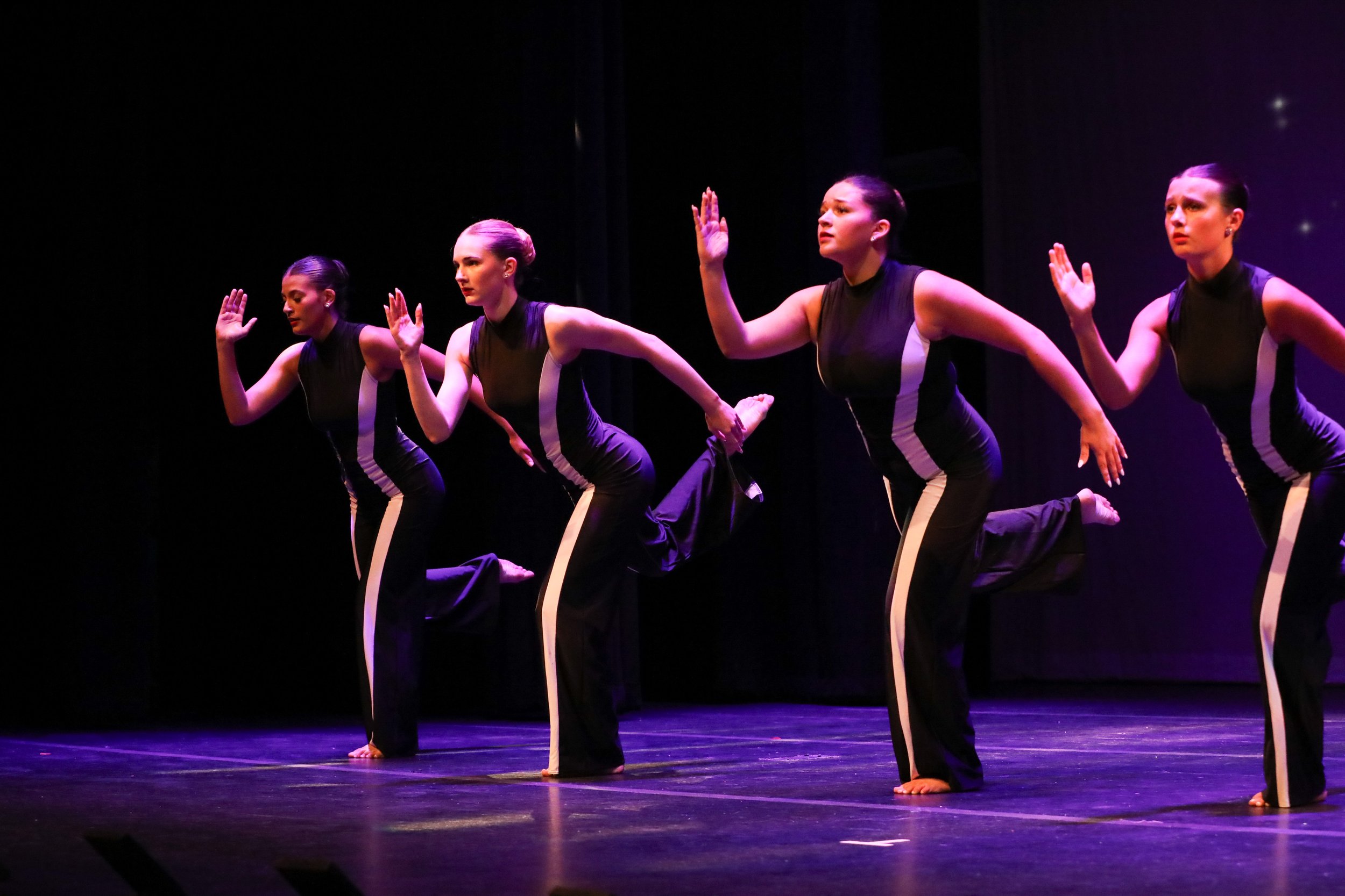 Four women in black dance costumes perform a synchronized dance on stage with purple lighting.
