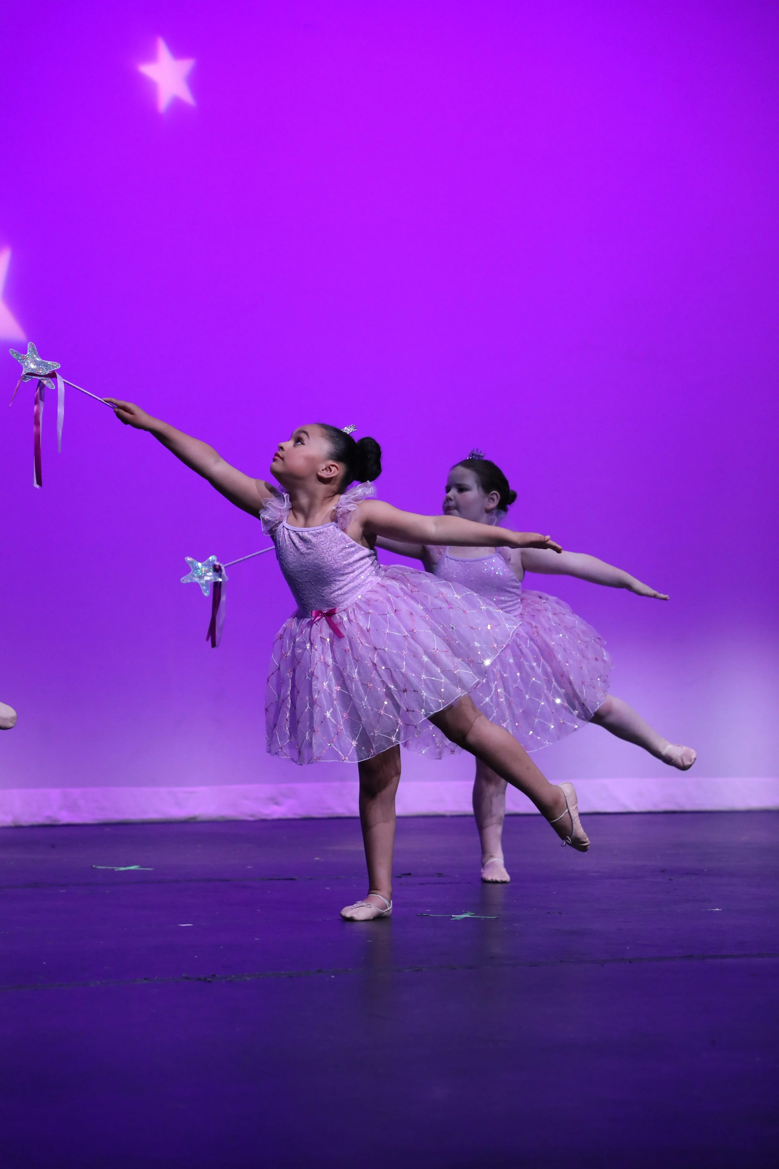 Two young ballet dancers in pink tutus and tiaras perform on stage against a purple background, holding star-shaped wands.