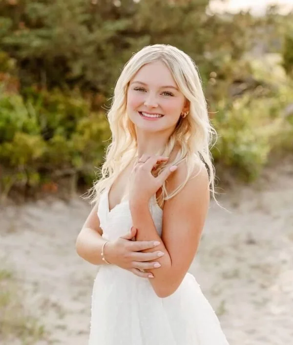 A young woman with long blonde hair wearing a white dress, smiling outdoors on a sunny day with greenery in the background.
