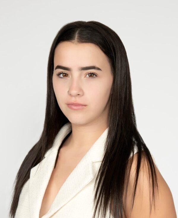 A young woman with long dark hair, fair skin, and a neutral expression, wearing a white sleeveless top, against a plain light background.