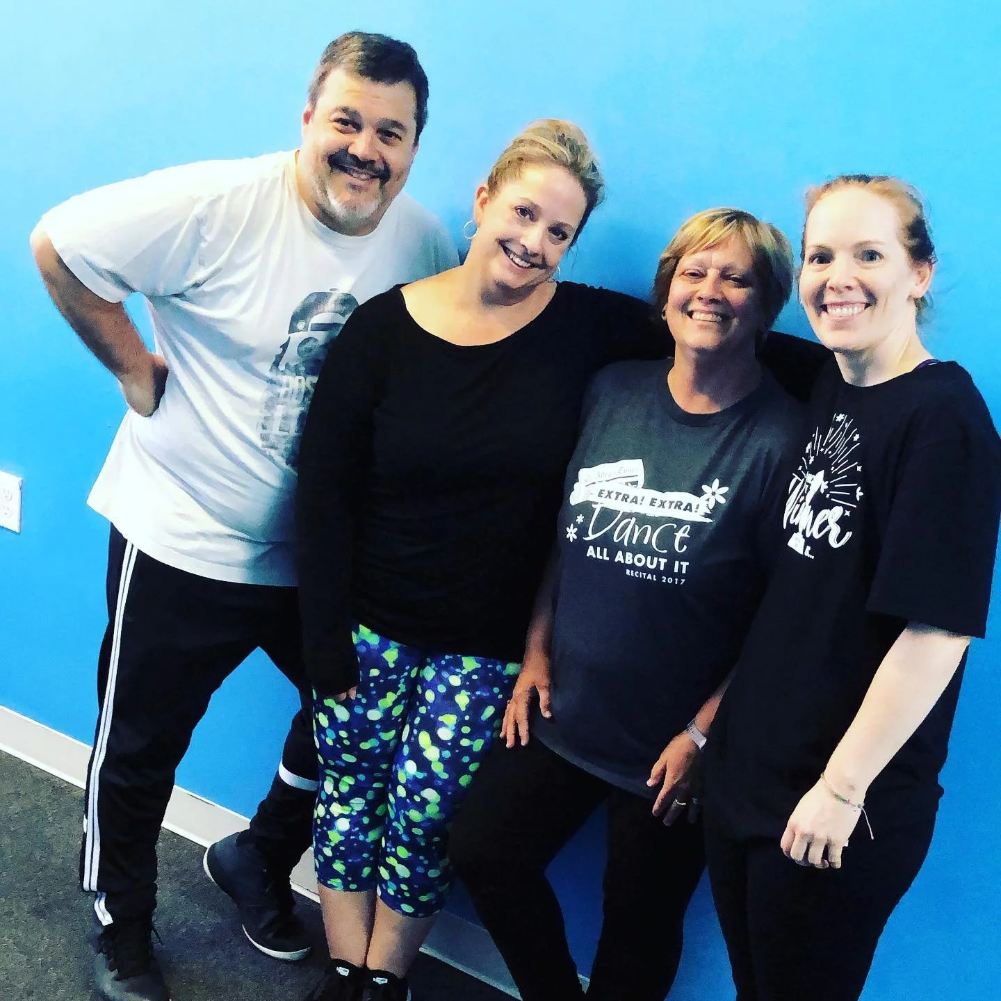 Four people standing against a blue wall, smiling at the camera. They are wearing casual clothes, with one person wearing a shirt that says 'Extra! Extra! Dance All About It'.