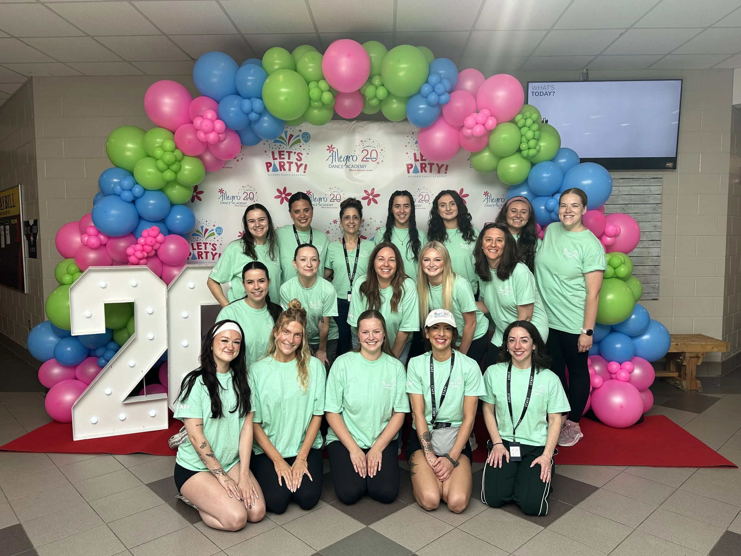 Group of smiling women in matching teal t-shirts gathered in front of a colorful balloon arch at a celebration, with a backdrop that reads 'Let's Party!' and 'Allegro Dance Academy.'