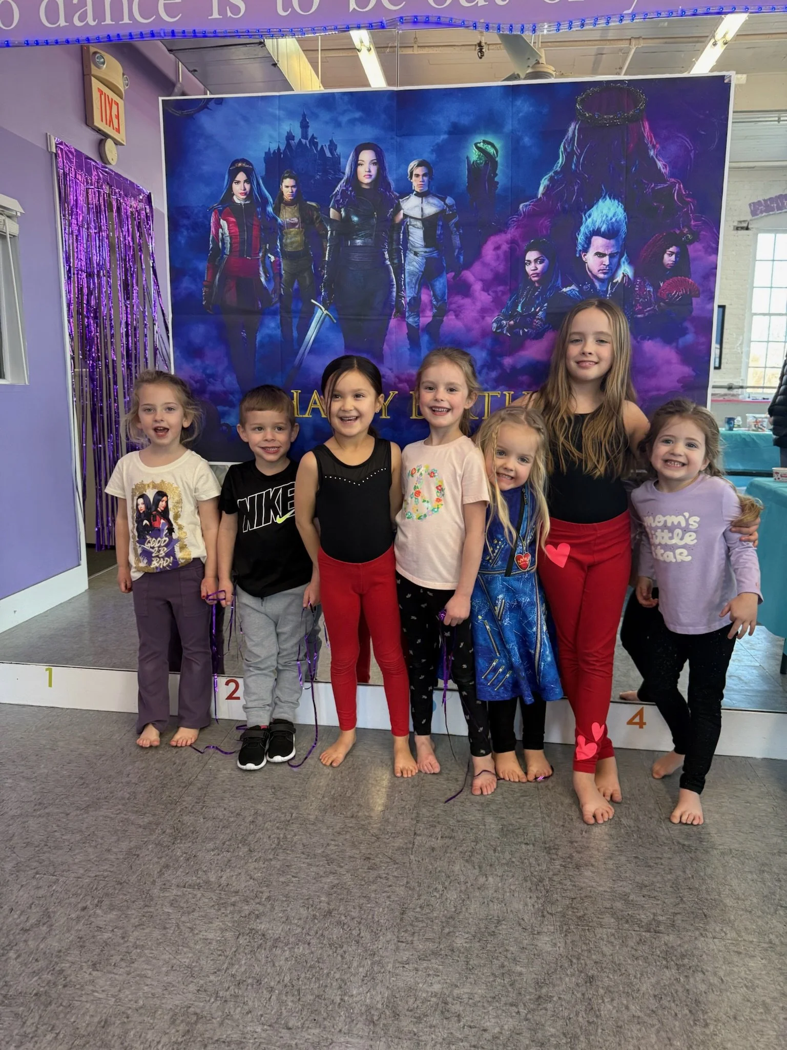 A group of seven young children standing barefoot in front of a large poster from the movie 'Harry Potter.' The children are smiling and appear to be in a celebratory mood, some wearing casual clothes and a few holding purple strings. The poster in the background features characters from the film with magical and fantasy imagery.