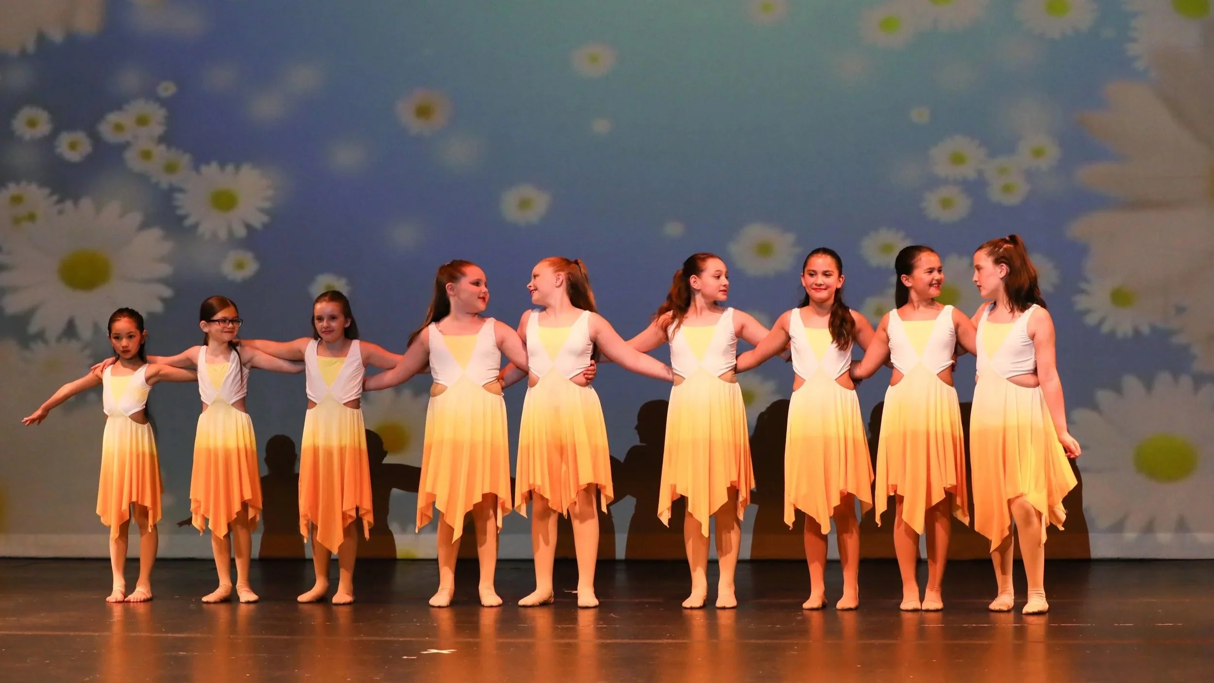 A group of nine young girls in matching yellow and white dresses, standing in a line on a stage, holding hands, with daisies and a blue sky background projected behind them.