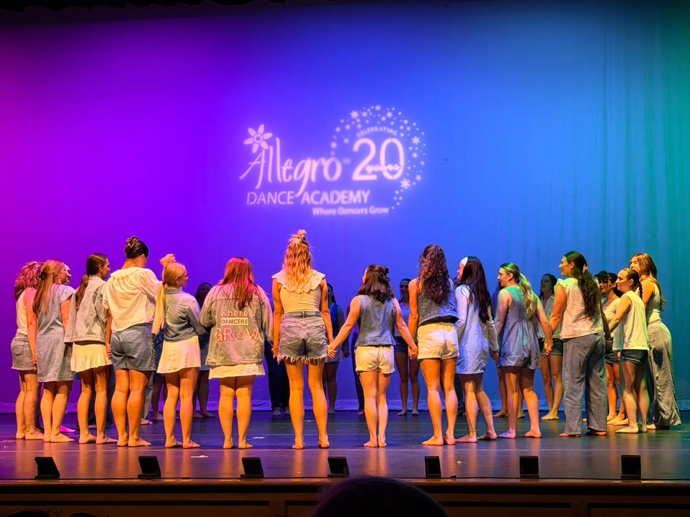 A group of dancers holding hands on stage during a performance at Allegro 20 Dance Academy's celebration. The stage is lit with colorful lighting, and the dancers are wearing casual dance attire.