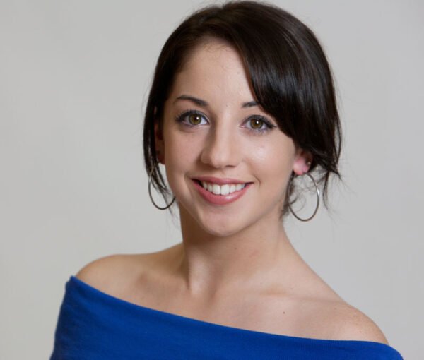 A woman with short dark hair wearing a blue off-the-shoulder top, smiling at the camera against a plain gray background.