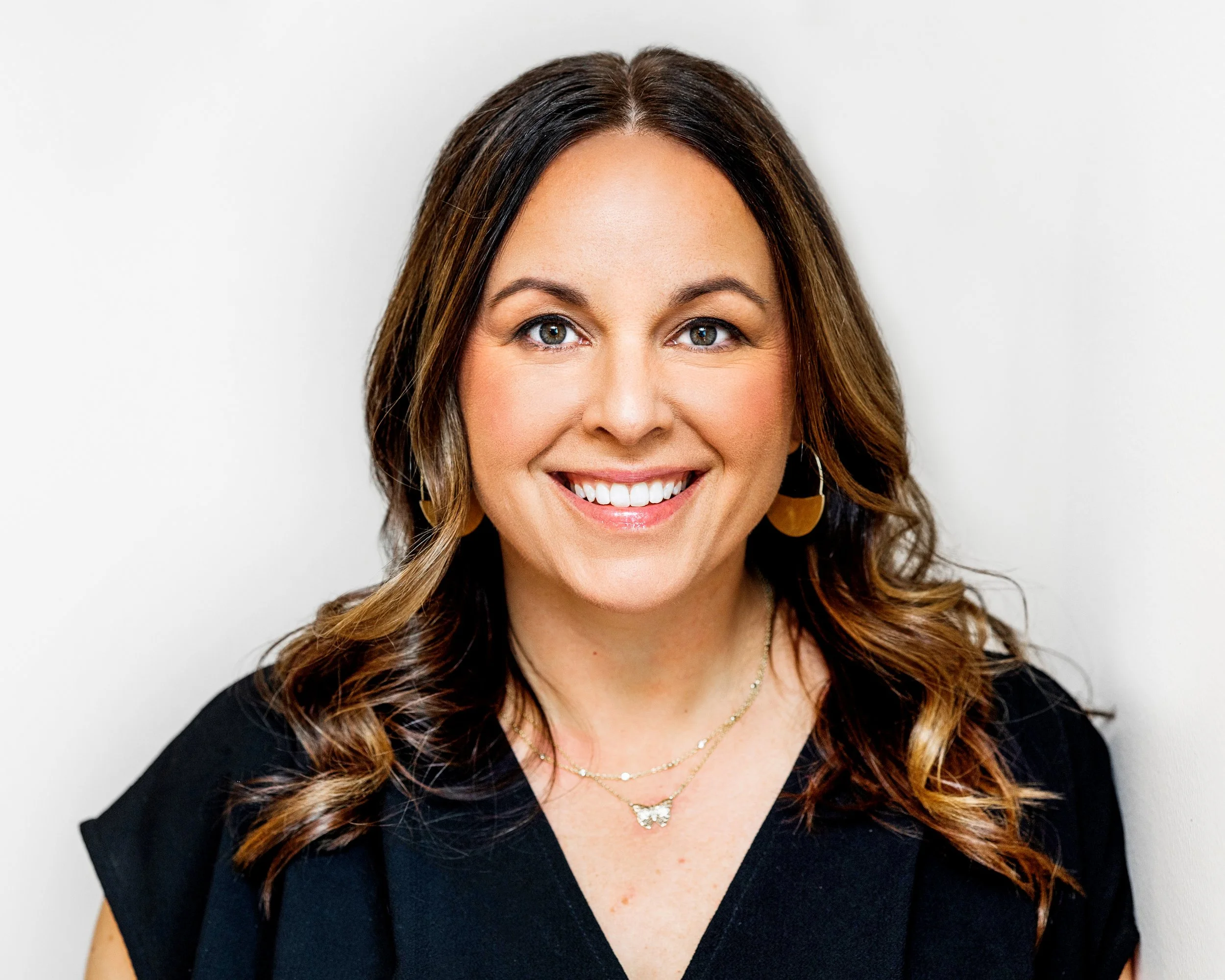 Close-up of a woman with long, wavy brown hair with highlights, smiling, wearing gold earrings and a butterfly-shaped pendant necklace, against a plain white background.