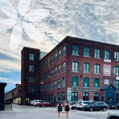 A historic red brick building with multiple floors and many windows, situated on a street with parked cars and two women walking in front.
