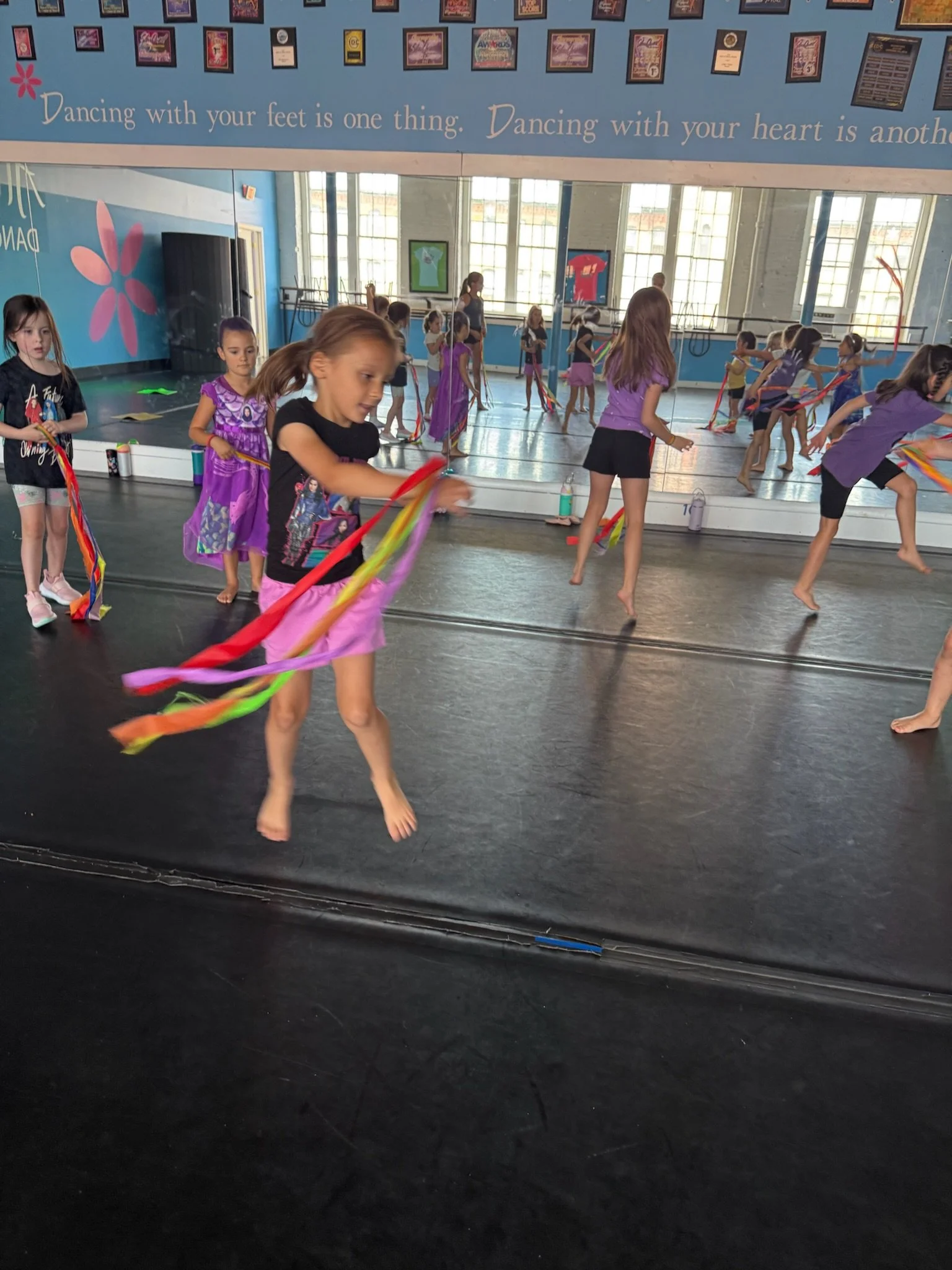 Young girls practicing dance with ribbons in a dance studio with large mirrors, blue walls, and framed pictures. Some girls are standing, others are in motion, barefoot, wearing casual dance attire.