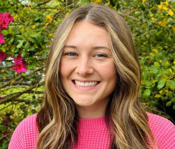 A young woman with long wavy brown hair, smiling, wearing a pink sweater, outdoors with green foliage and pink flowers in the background.