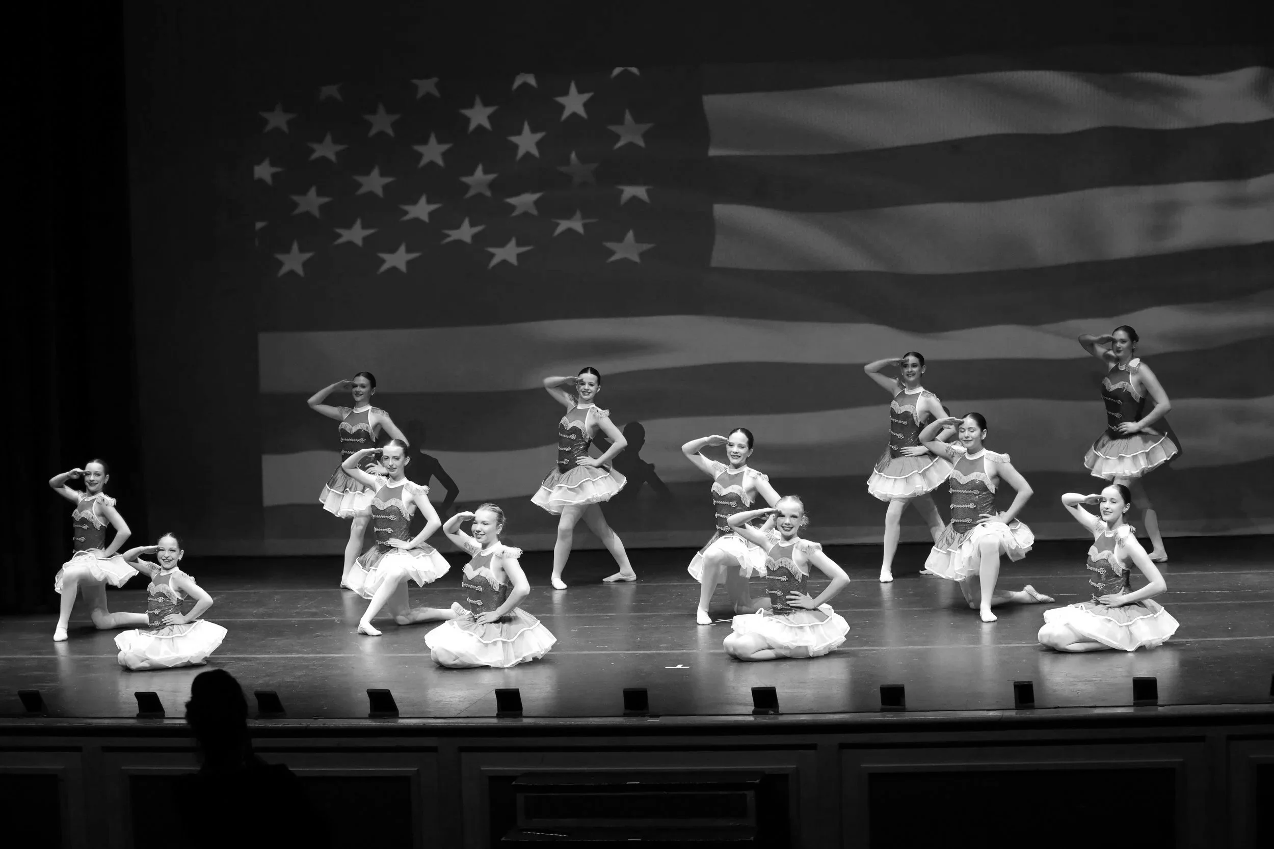 Black and white photo of young ballet dancers performing on stage with an American flag projected in the background.