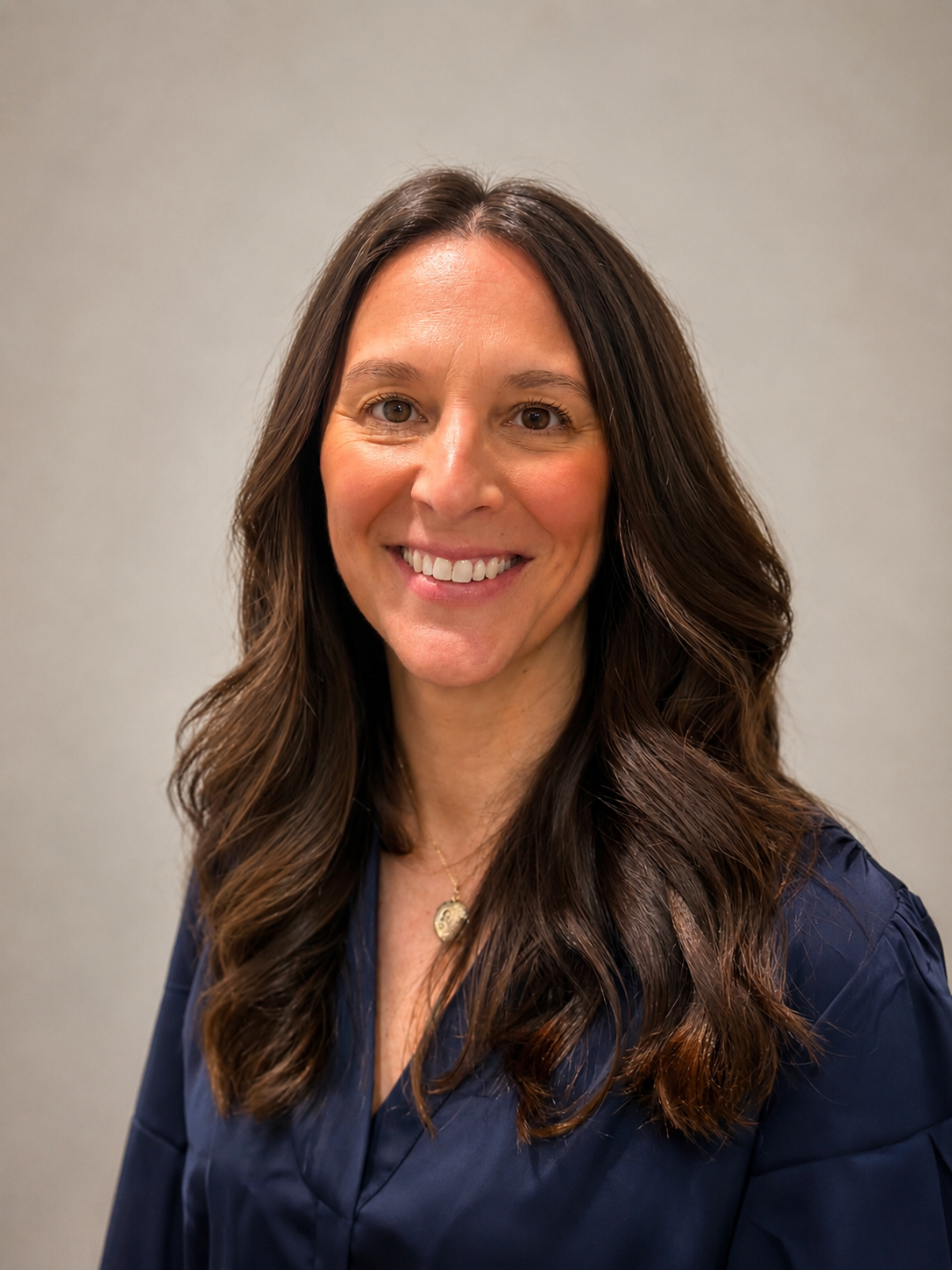 A woman with long brown wavy hair smiling in front of a plain gray background.