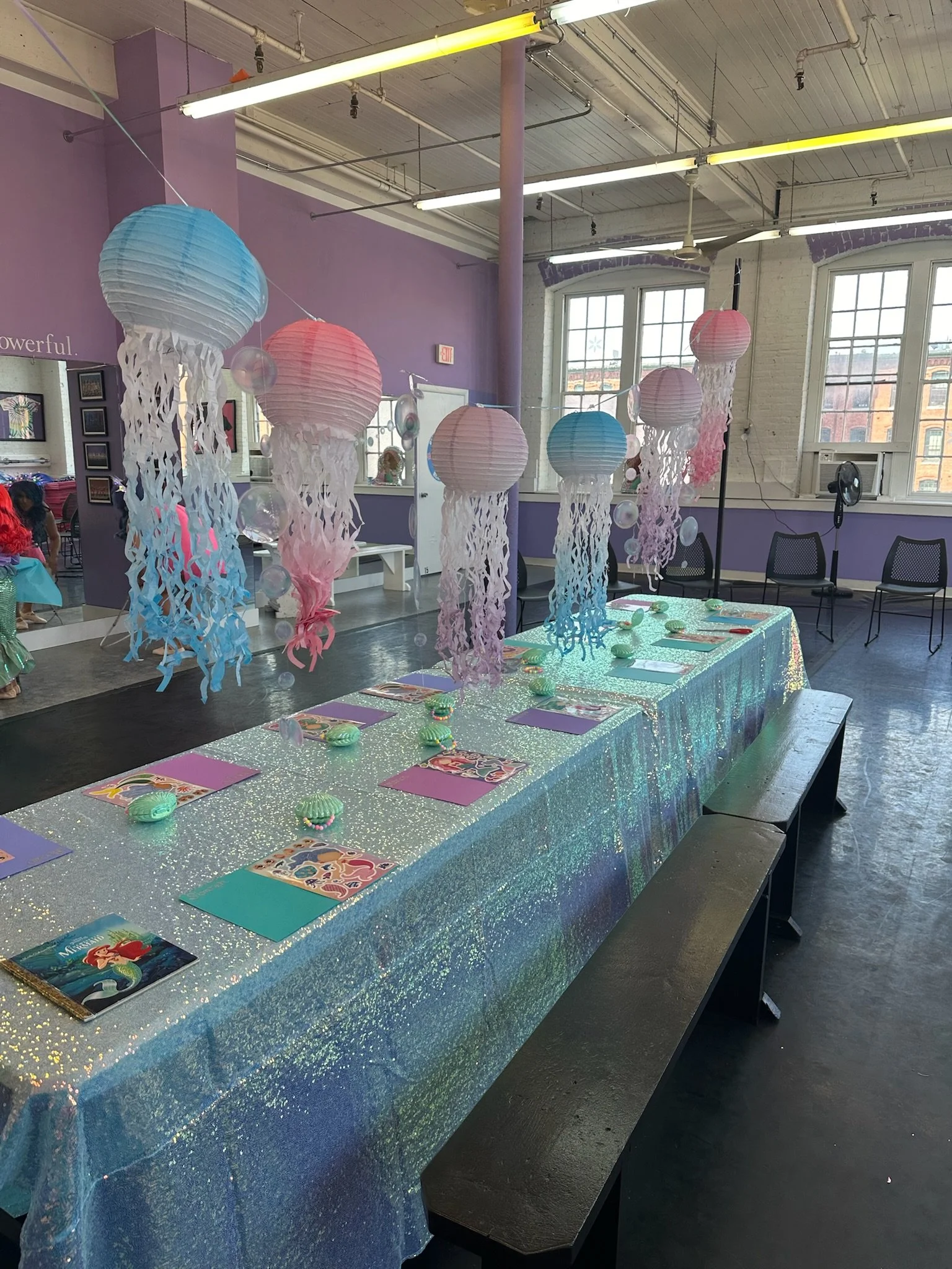 Decorative table covered in sparkly blue fabric with seashells, mermaid-themed coloring pages, and pink, blue, and purple hanging lanterns with paper streamers inside a spacious room with large windows, purple walls, and fluorescent lighting.