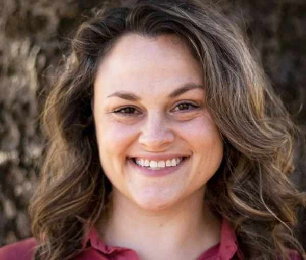 A young woman with curly brown hair smiling at the camera outdoors.