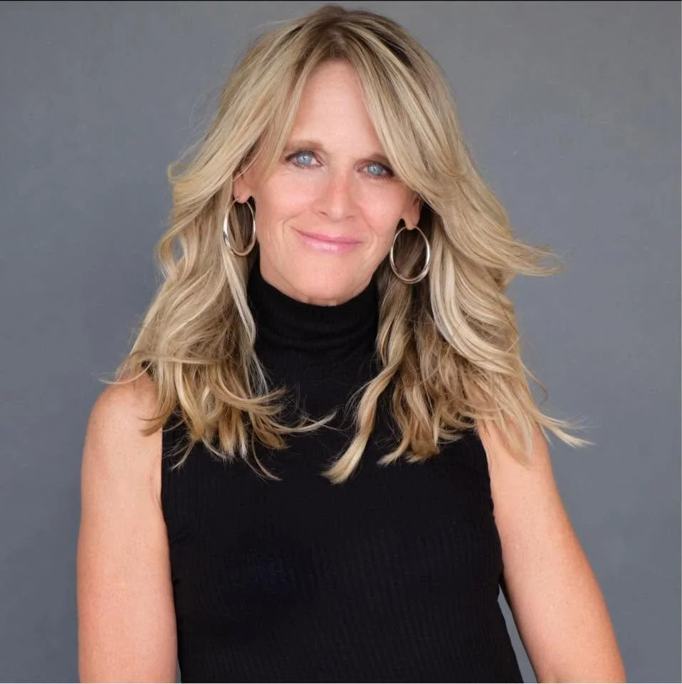 Woman with long blonde hair, blue eyes, wearing a sleeveless black turtleneck sweater and hoop earrings, smiling against a gray background.