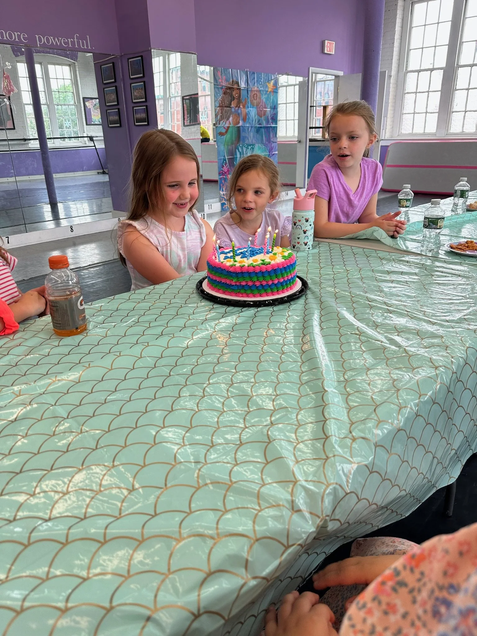 Three young girls sitting at a table celebrating a birthday with a colorful frosted cake with lit candles, surrounded by water bottles and party supplies, in a dance studio with purple walls and large windows.