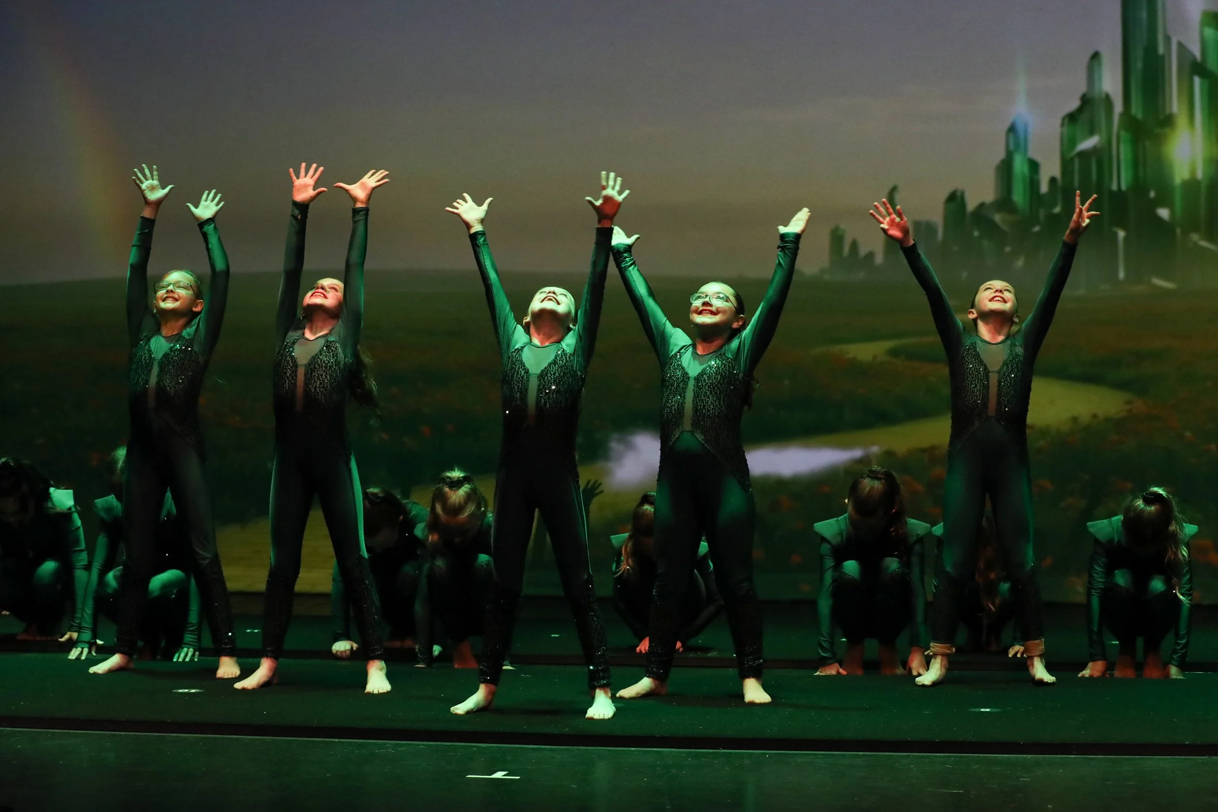 Group of young girls performing dance on stage, dressed in black costumes with sheer sleeves, with some standing and some kneeling, under a backdrop of a cityscape projection.