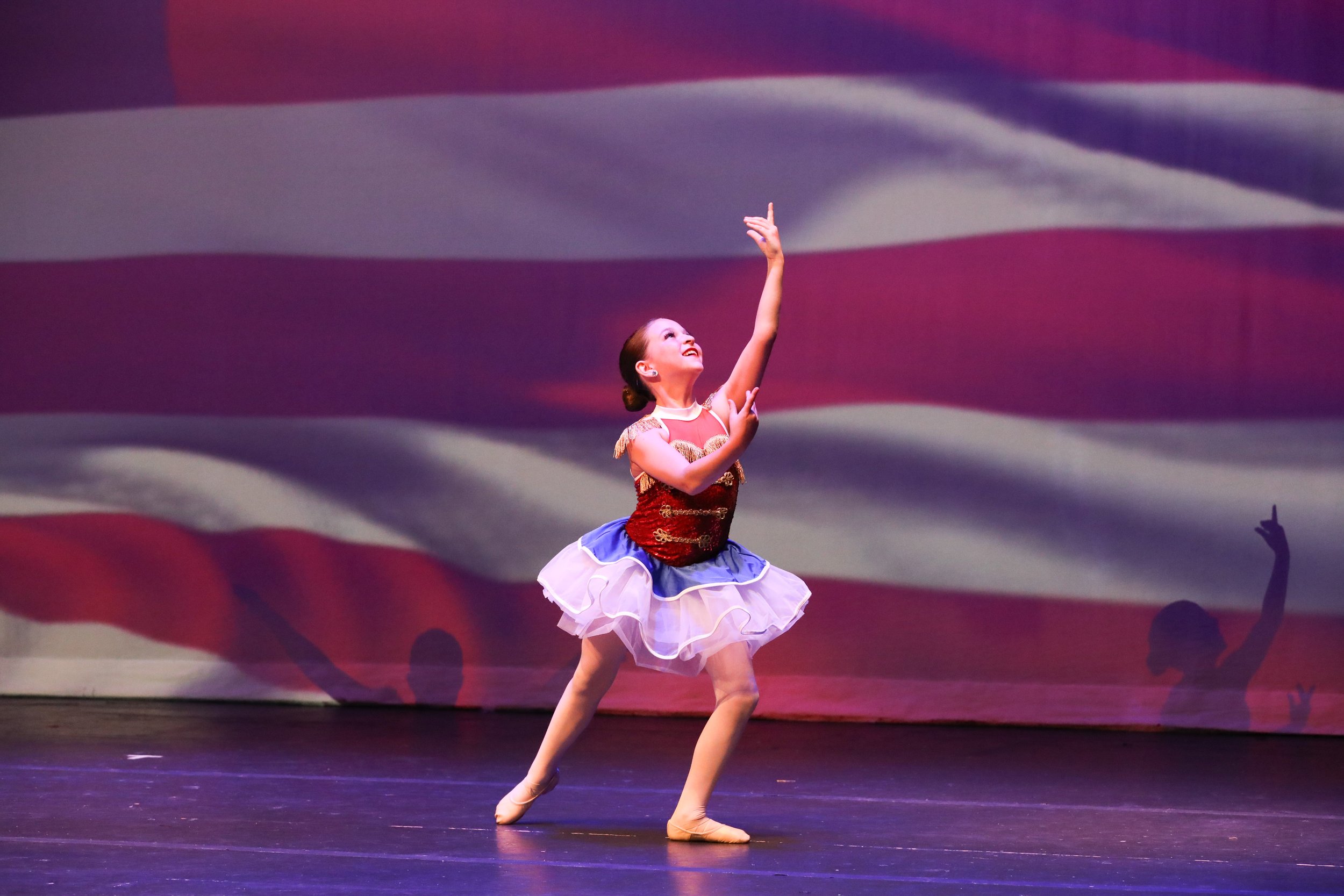 A young girl performing a ballet dance on stage, wearing a red and gold top with a white tutu and beige ballet shoes, with an American flag backdrop projecting in the background.