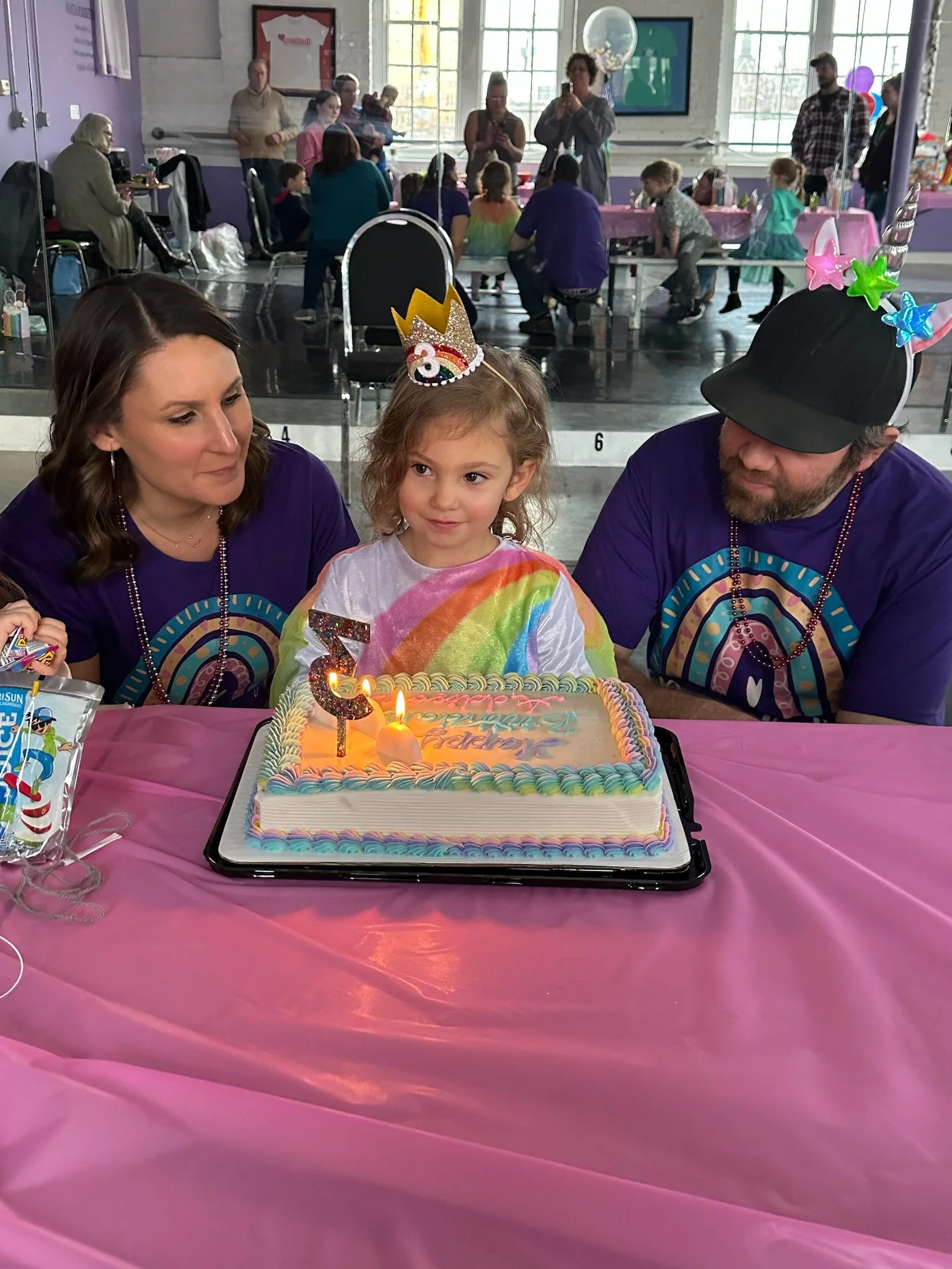 A young girl celebrating her birthday with a cake, wearing a rainbow-colored shirt and a birthday crown. She is sitting at a table with her parents, who are wearing matching rainbow-themed shirts. The cake is decorated with colorful frosting and has candles on it. In the background, there is a party setting with other people, children, balloons, and decorations.