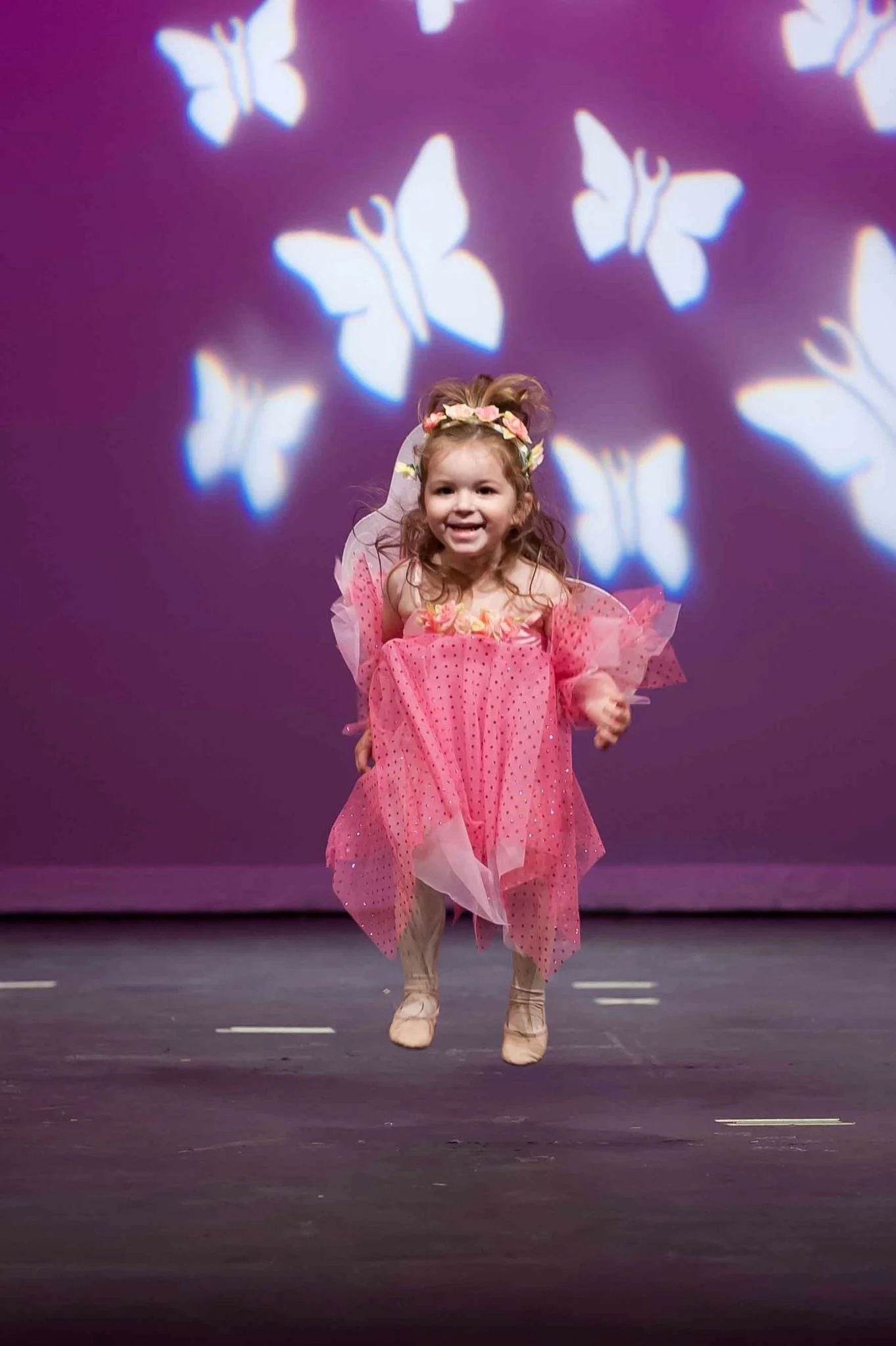 Young girl in a pink fairy costume with wings, a floral headband, and ballet shoes, smiling on stage with illuminated butterfly shapes projected on a purple background.