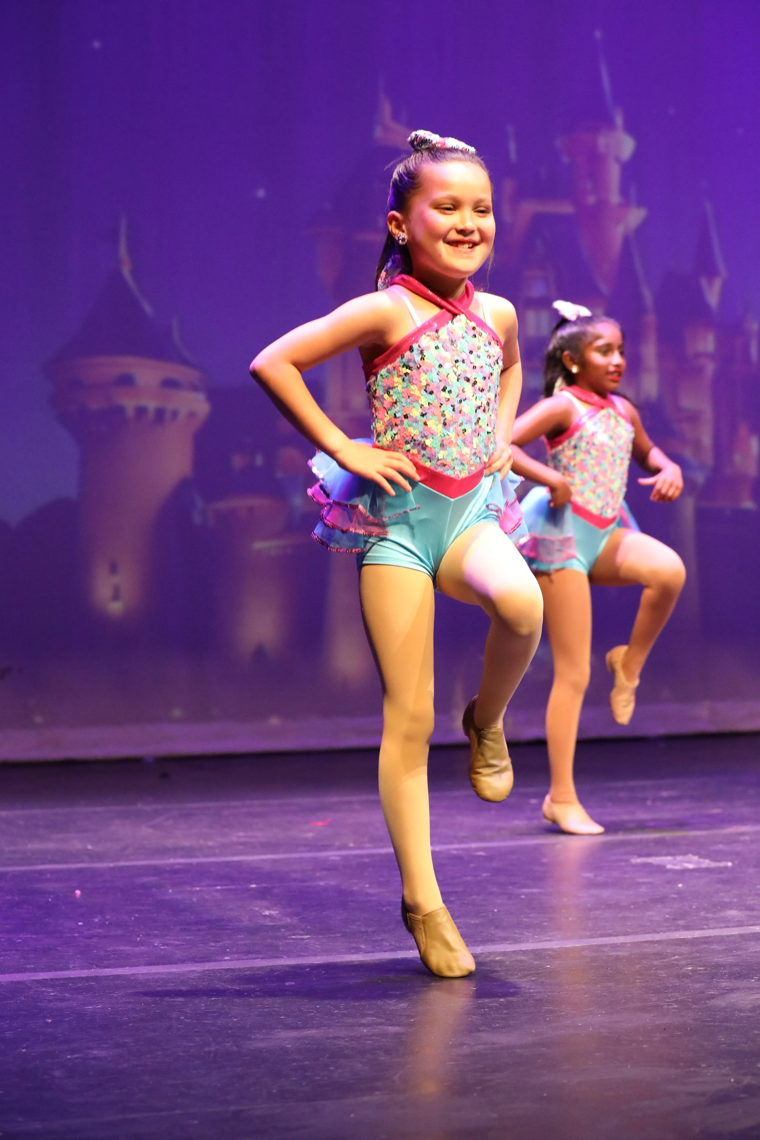 Young girls in colorful dance costumes performing on stage with a castle backdrop.