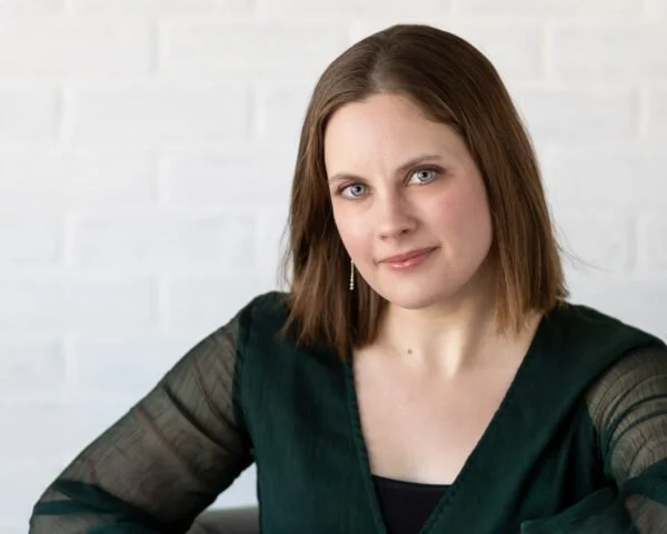 A woman with shoulder-length brown hair and blue eyes, wearing a black shirt and a dark green blouse with sheer sleeves, sitting in front of a white brick wall.