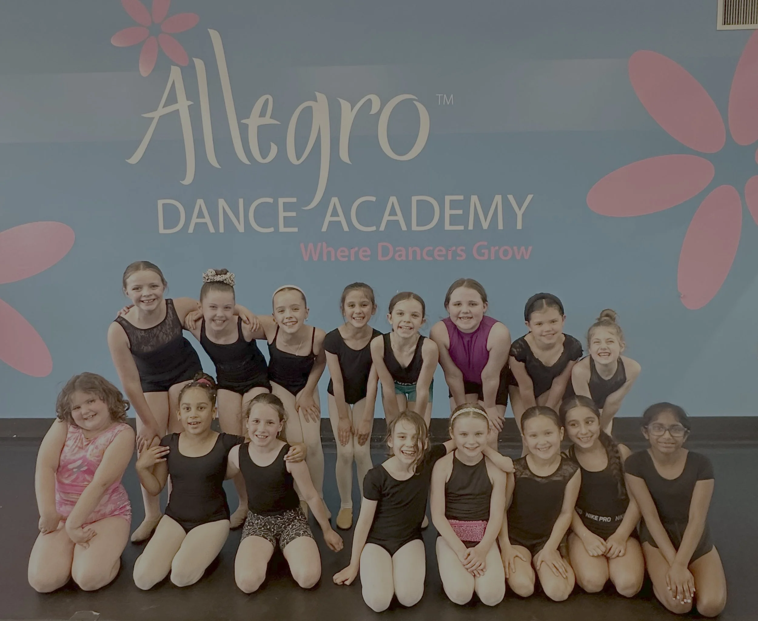 Group of young girls smiling and posing for a photo at a dance academy. They are wearing dance attire, with the background displaying the name 'Allegro Dance Academy' and a tagline 'Where Dancers Grow'.