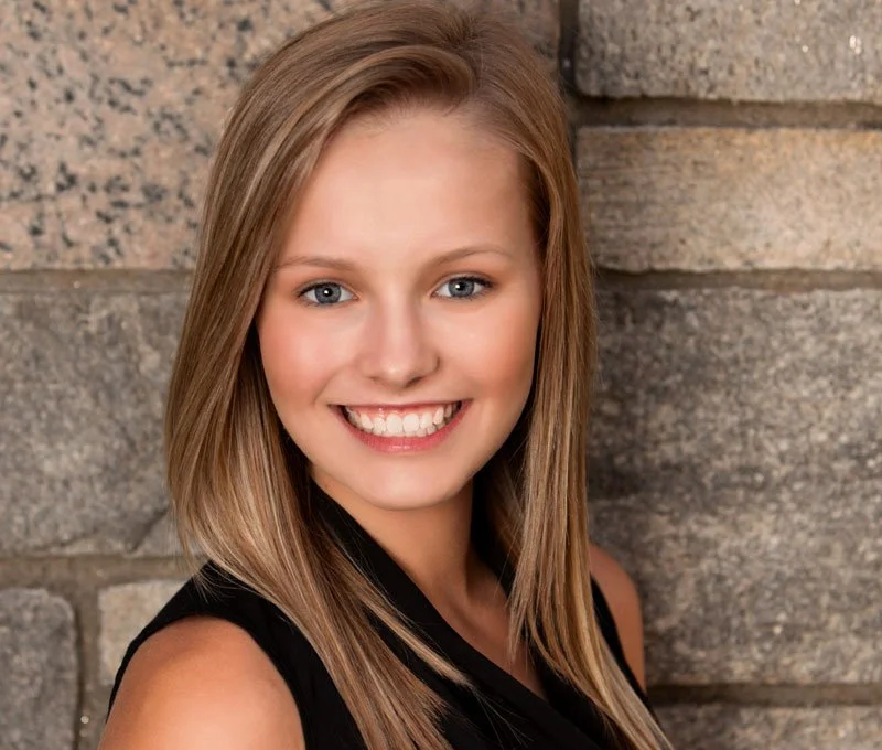 A young woman with blonde hair and blue eyes smiling at the camera, wearing a sleeveless black top, standing against a brick wall.