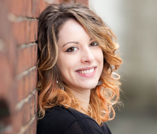 A young woman with curly, shoulder-length hair leaning against a brick wall and smiling at the camera.
