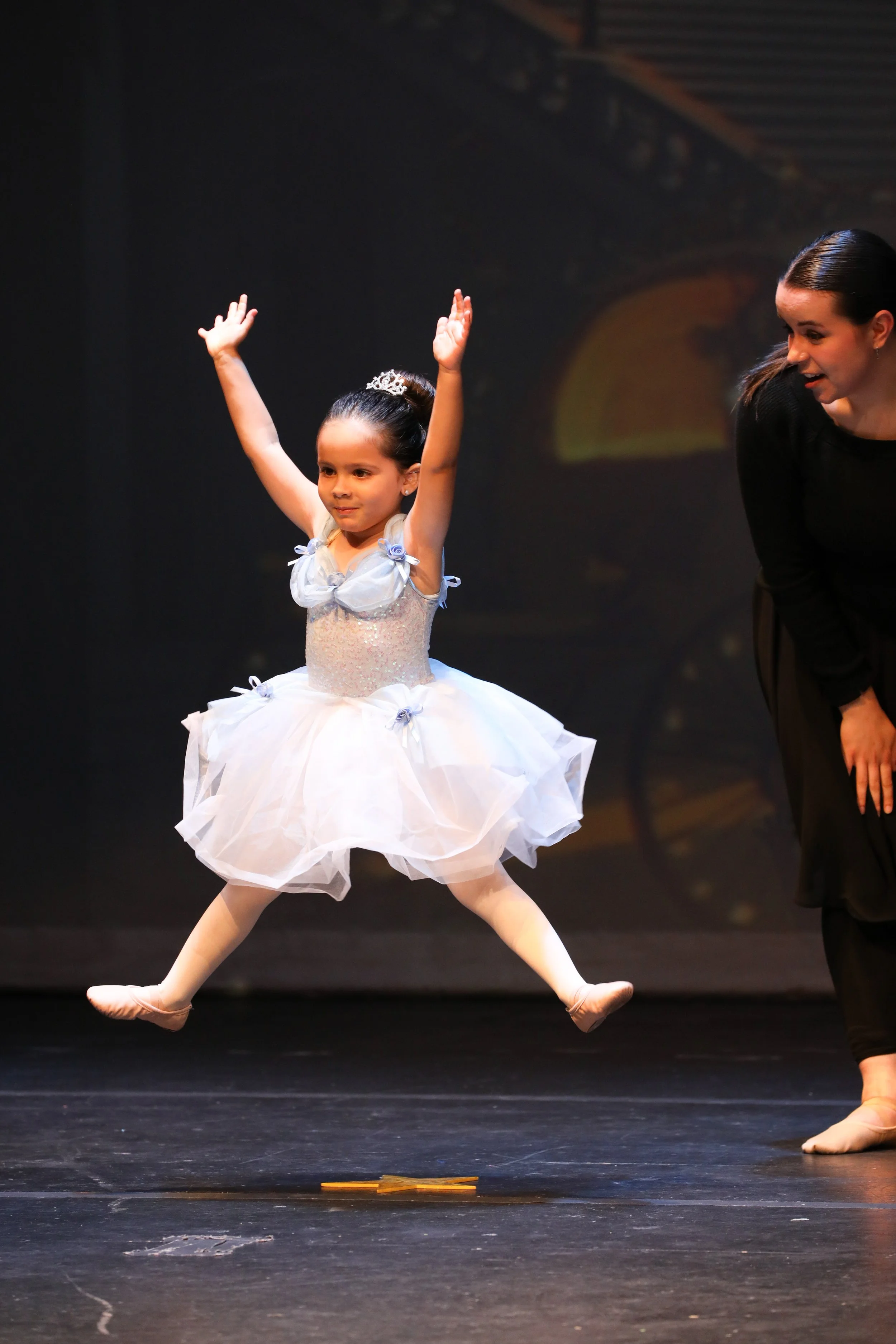 A young girl dressed in a white ballet costume performing a ballet dance on stage, with an adult woman nearby, possibly her teacher or a fellow performer, during a dance recital.