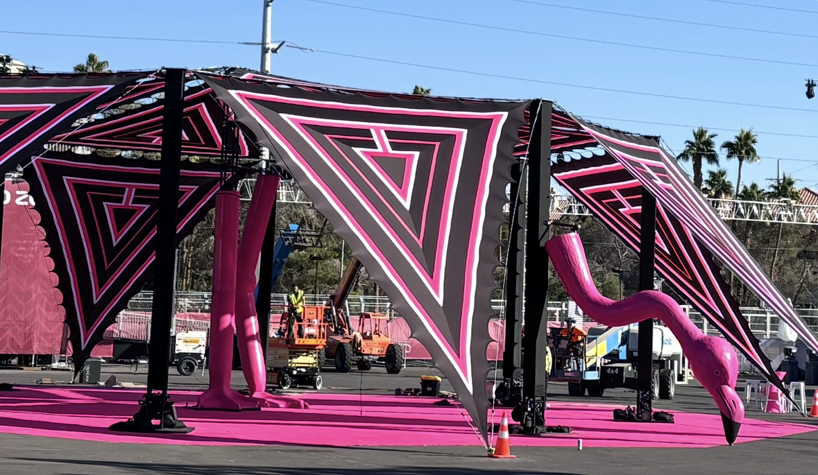 Stage set with pink and black geometric patterned tents, a giant pink flamingo decoration, and construction workers preparing the area.