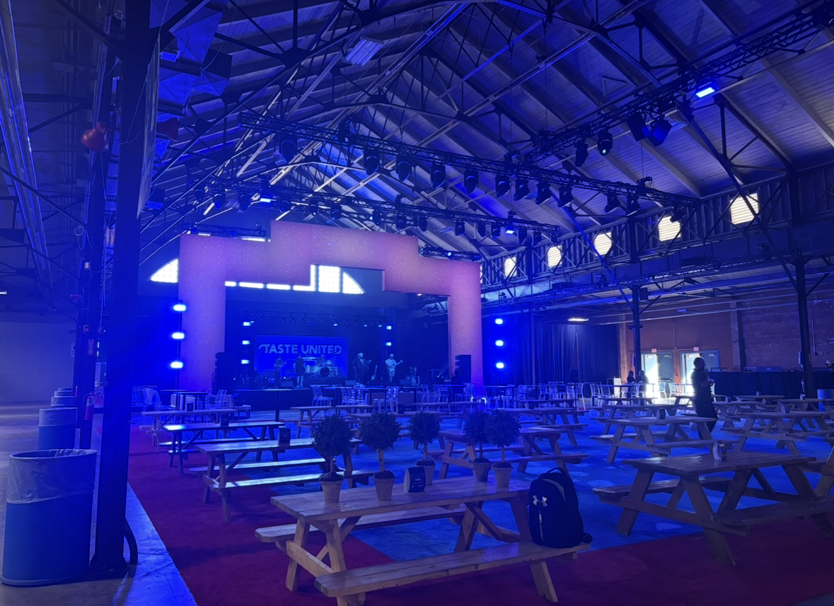 An indoor event space with a stage, musical band, blue lighting, picnic tables with potted plants, and a few people preparing for a performance.