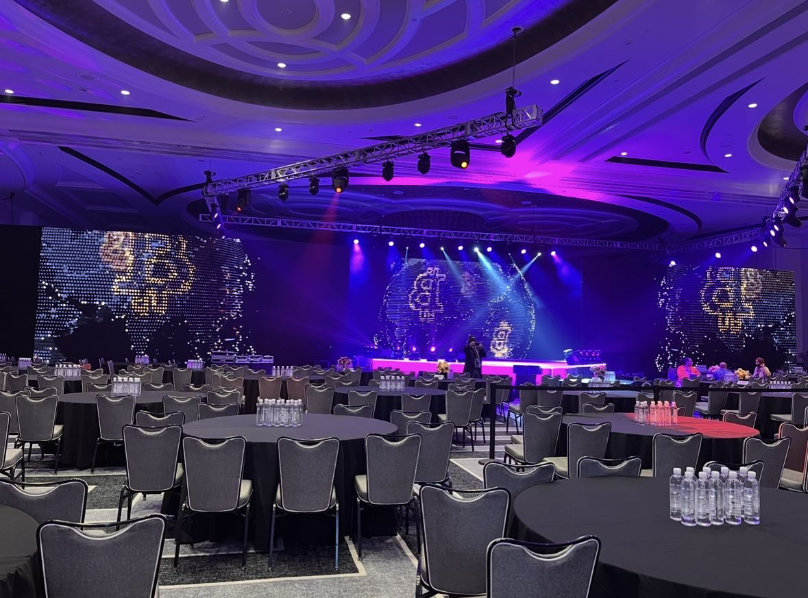 A large event hall set for a formal gathering with round tables, black tablecloths, bottled water on each table, and a stage with colorful LED screens and lighting effects, ready for a presentation or performance.