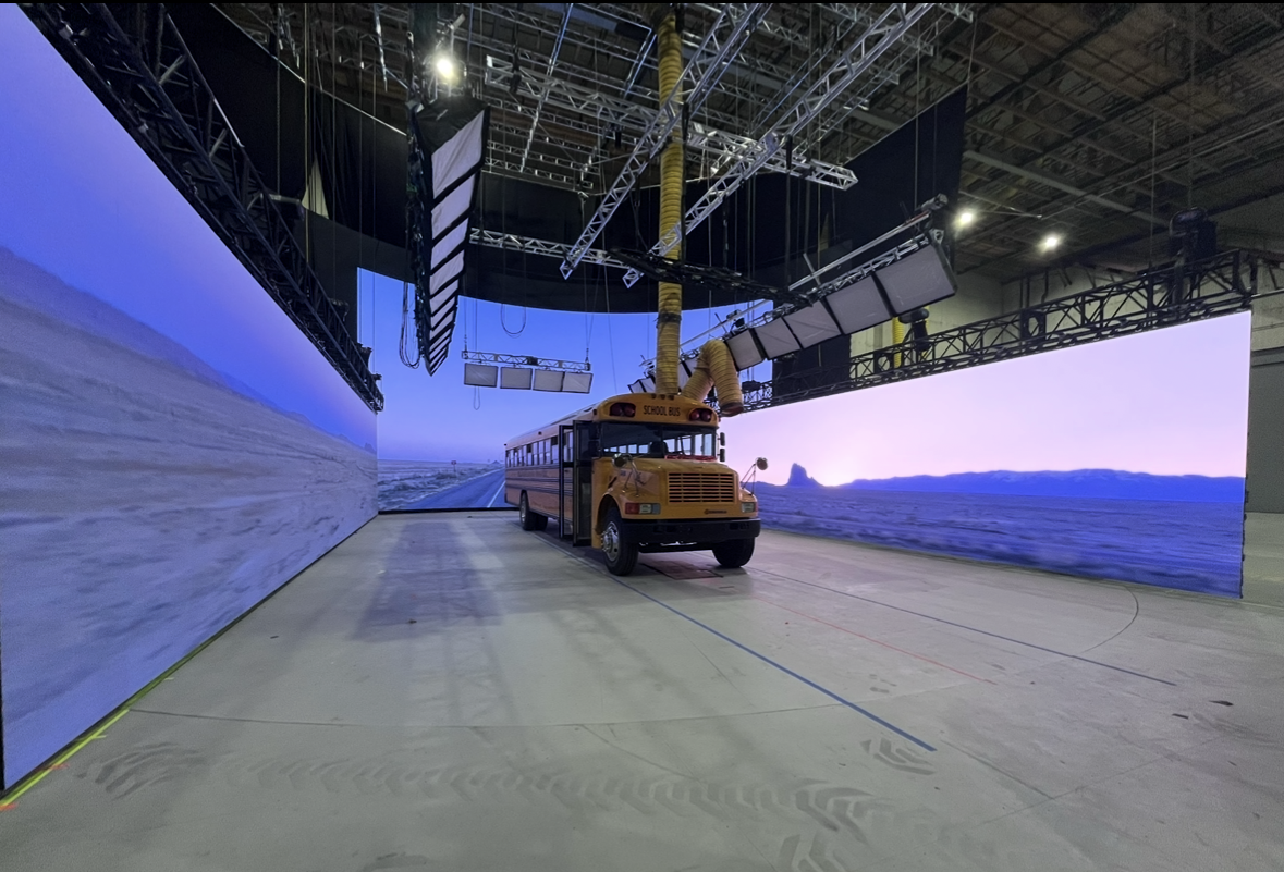 Indoor studio setup with large digital screens, a yellow school bus model on the floor, and stage lighting and equipment hanging from a metal truss structure overhead.