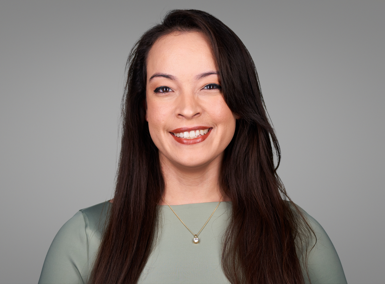 Portrait of a smiling woman with long dark hair, wearing a light green top and a delicate gold necklace with a small pendant, against a plain gray background.