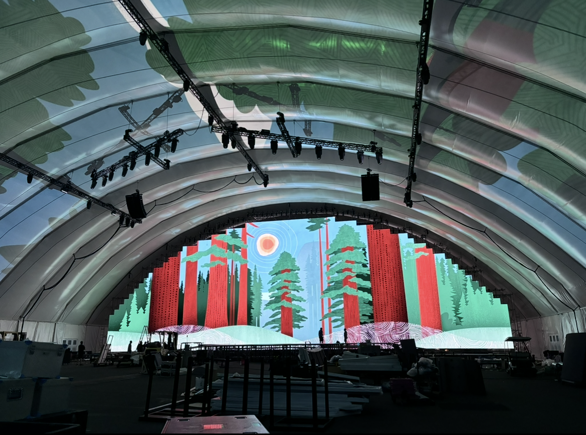 A large stage with a digital forest landscape backdrop, featuring tall trees, a sun, and stylized natural scenery, inside an arched tent.