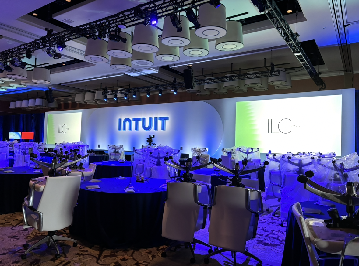 Conference room set up with round tables, white chairs, and stage with large illuminated signs displaying 'INTUIT' and 'ILC 25', decorated with draped fabric and robotic equipment.