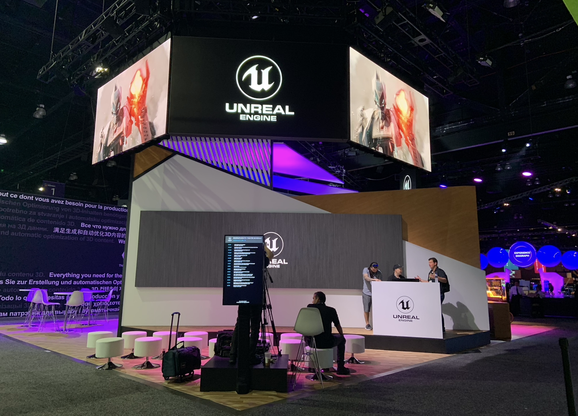 Expo booth for Unreal Engine with large digital screens displaying game characters, a reception desk, seating areas, and people working at the booth.