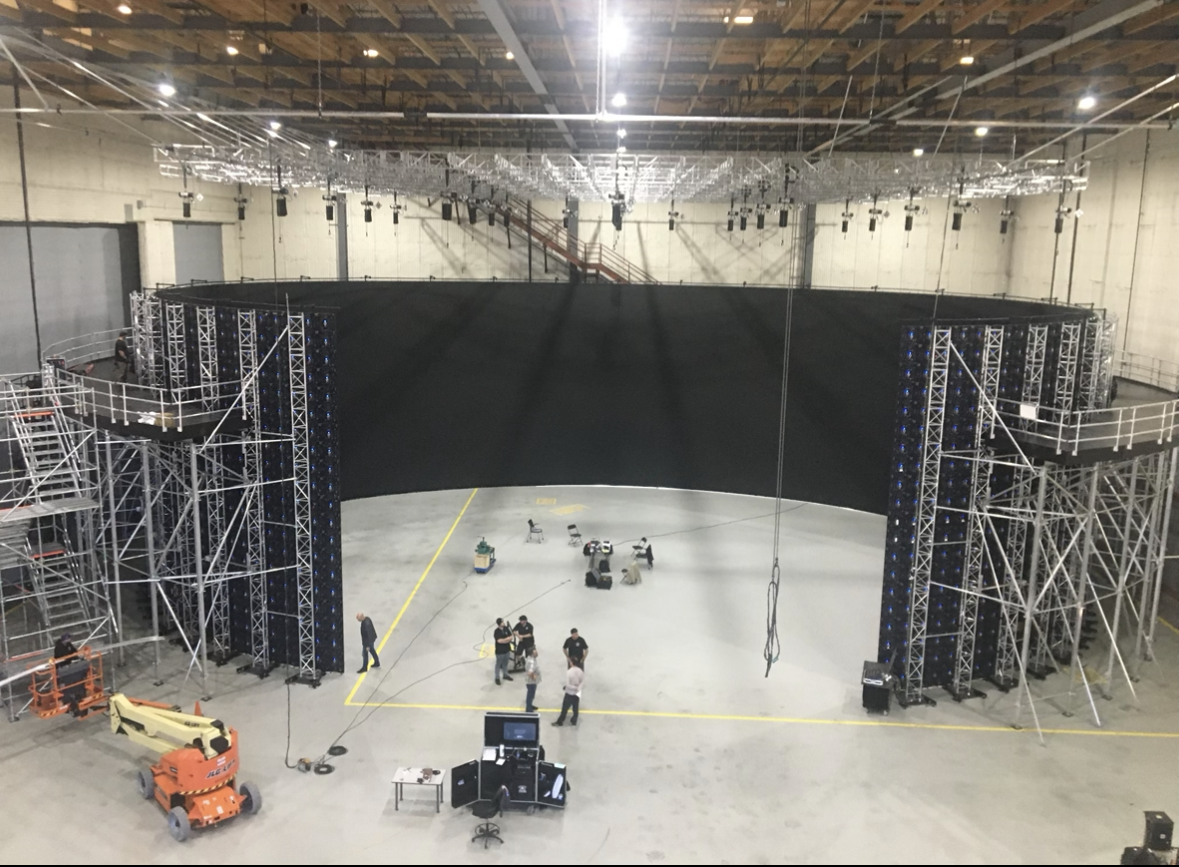 Workers setting up a large black curved LED screen inside an industrial warehouse with scaffolding and lighting equipment.