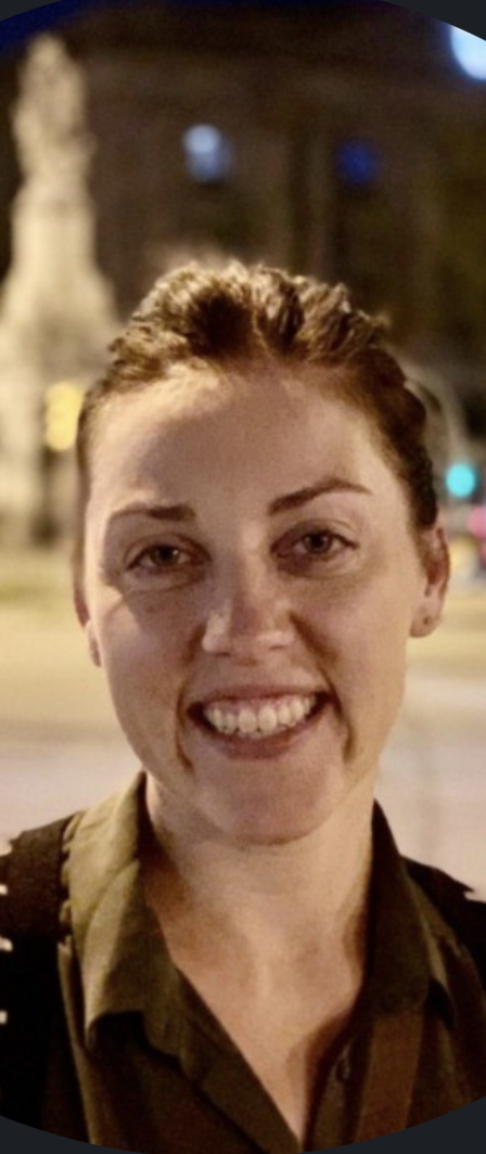 A woman with red hair smiling outdoors at night, with blurred city lights and buildings in the background.