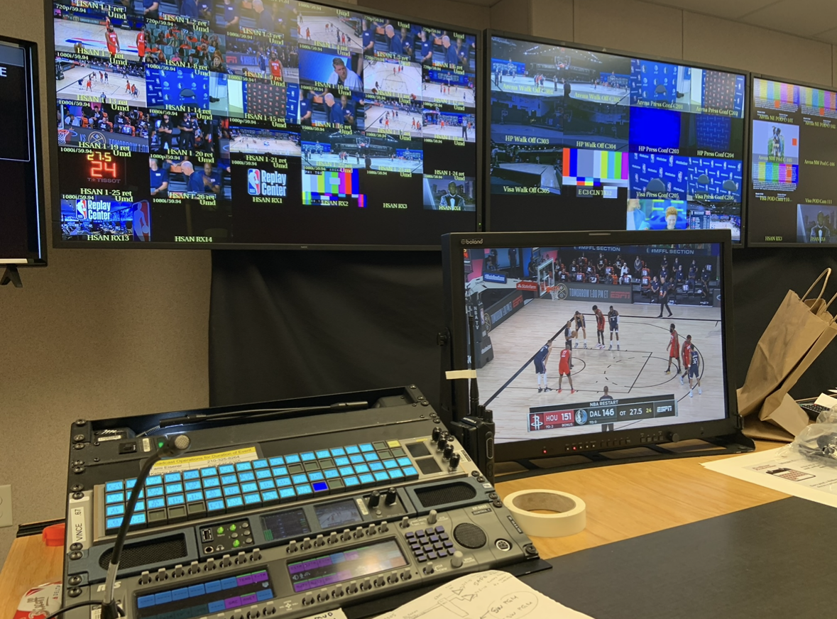 Control room with multiple monitors showing live sports broadcasts and a basketball game on a computer monitor. There is audio or video equipment on the desk, notebooks, and other items.