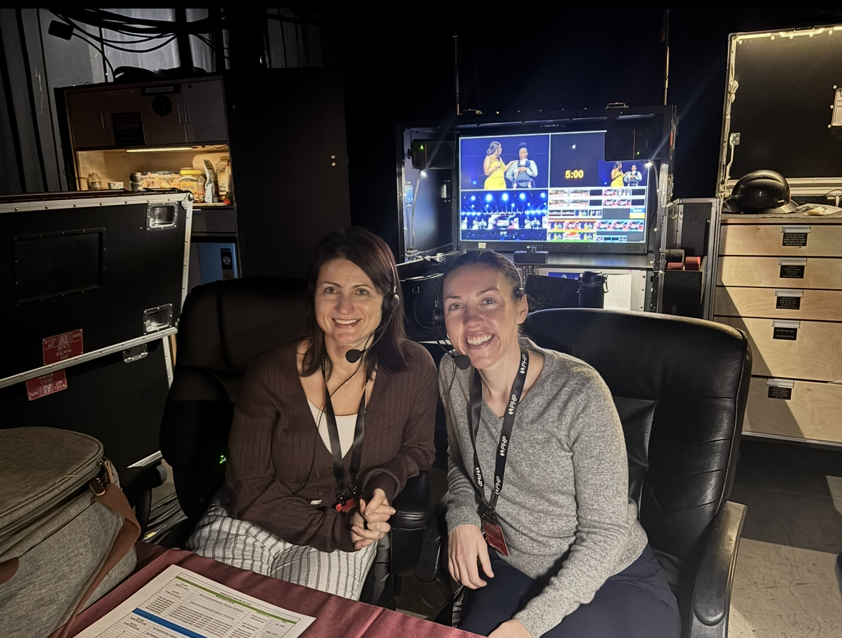 Two women wearing headsets and smiling at the camera in a television control room with monitors and equipment.