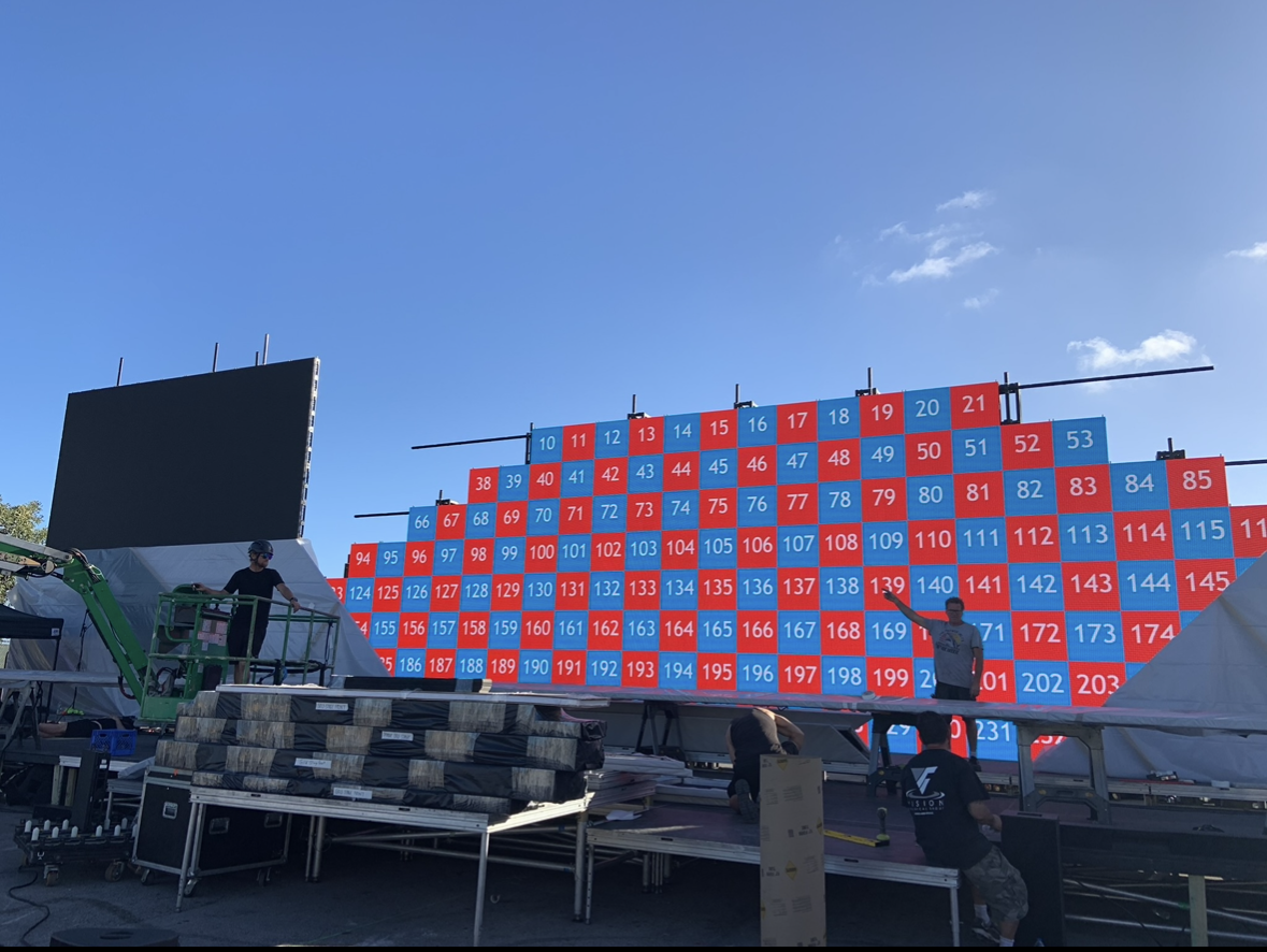 Preparations for a large LED display, with a grid of numbered red and blue sections, on a stage outdoors under a clear blue sky.