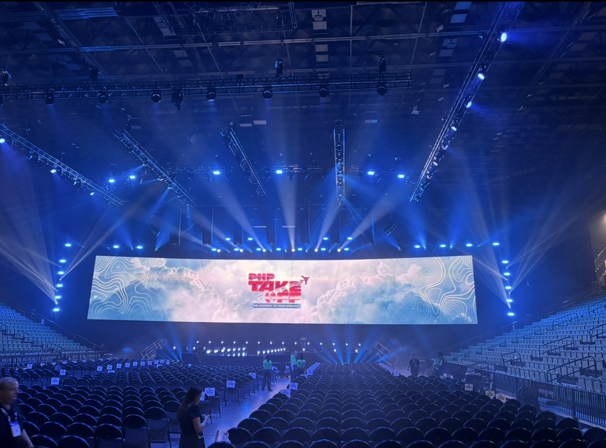 Empty auditorium with rows of chairs and a large stage featuring an LED screen displaying 'PHP TAKE OFF' with clouds in the background and concert lighting.