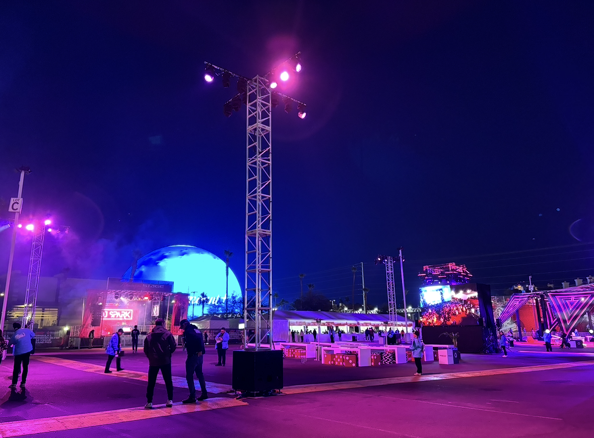 Night scene at an outdoor music event with colorful stage lights, large screens, and people walking around.